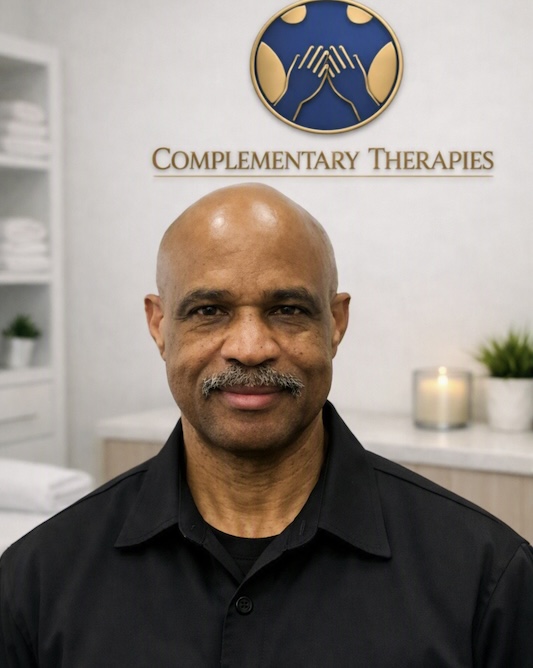 Portrait of a bald man with a mustache in a black shirt, standing inside a complementary therapies clinic with a logo and 'Complementary Therapies' sign behind him.