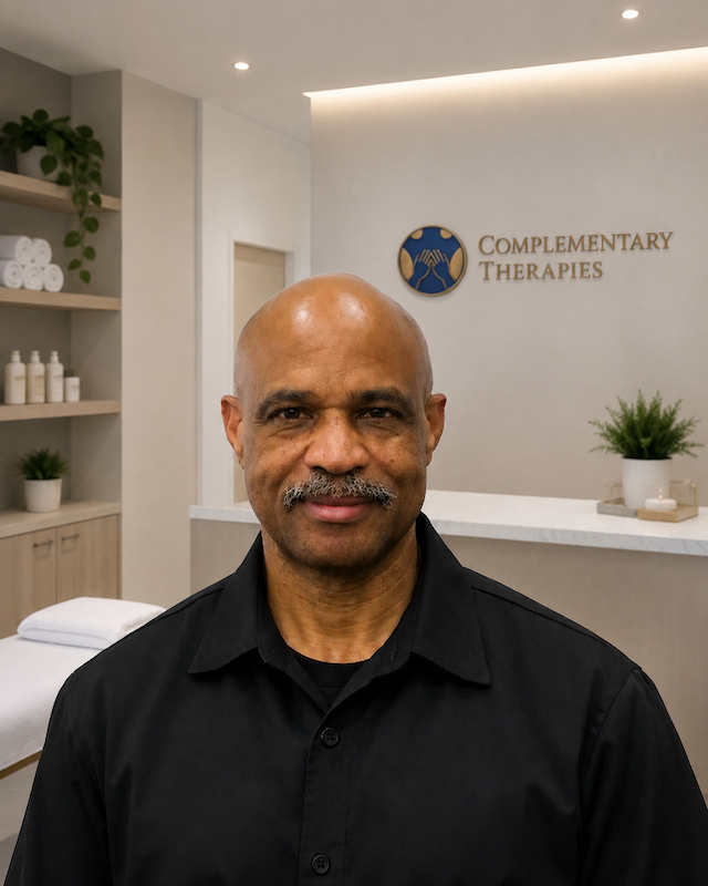 Smiling man in a black shirt stands in a spa reception area with a sign reading 'Complementary Therapies' on the wall.